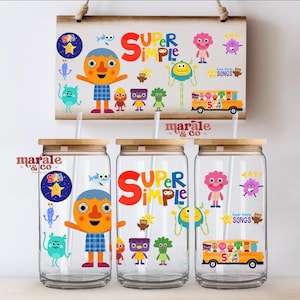 Può includere: Tre bicchieri di vetro trasparente con coperchi in bambù e cannucce, ciascuno decorato con personaggi dei cartoni animati colorati e le parole "SUPER SIMPLE". Un cartello abbinato è appeso sopra.