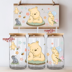 May include: Three glass tumblers with bamboo lids and straws, featuring Winnie the Pooh illustrations. The design includes Pooh Bear, Eeyore, Piglet, bees, and flowers. A matching wooden sign hangs above the tumblers, also decorated with the same characters.