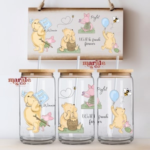 Può includere: Tre bicchieri in vetro trasparente con coperchi in bambù e cannucce, decorati con illustrazioni di Winnie the Pooh e Pimpi. I disegni includono vasi di miele, palloncini e api, con il testo We'll be friends forever. Un cartello rettangolare in legno abbinato è esposto sopra.