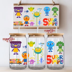 Può includere: Tre bicchieri di vetro trasparente con coperchi in legno e cannucce, ciascuno decorato con personaggi di cartoni animati colorati. Un cartello di legno sopra mostra personaggi simili e le parole "SUPER SIMPLE".