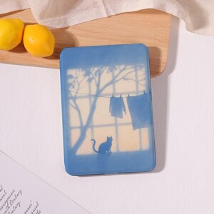 May include: A blue rectangular case featuring a silhouette of a cat, clothes on a clothesline, and a tree against a light background. The case is on a wooden surface with two yellow lemons.