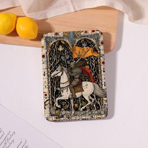 May include: A rectangular decorative plaque featuring a knight in armor riding a white horse. The knight holds a yellow flag and wears a red cape. The background depicts a dark forest scene framed by a stained glass style border with floral motifs.