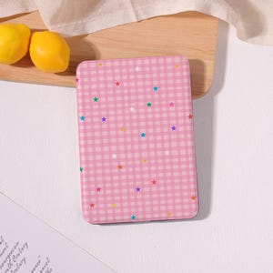 May include: A rectangular pink phone case with a gingham pattern and colorful star accents. The case is on a wooden surface with two yellow lemons nearby. The design is playful and bright.