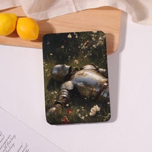 May include: Rectangular coaster featuring a painting of a knight in full armor lying in a field of wildflowers. The knight's silver armor contrasts with the green grass and colorful flowers. Two yellow lemons are visible in the background.