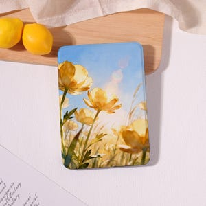 May include: A rectangular e-reader case featuring a printed illustration of yellow buttercup flowers in a field under a bright blue sky with a sun flare. The design shows green stems and leaves amidst the golden petals and tall grasses.