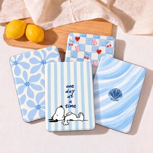 May include: Four rectangular cases with blue and white designs. One features a floral pattern, one a blue and white checkerboard with cherries and bears, one a blue striped design with Snoopy and the text one day at a time, and one a seashell.
