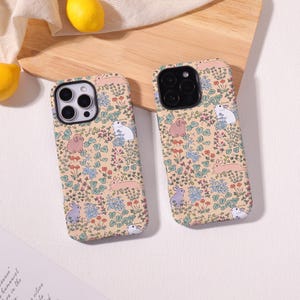 May include: Two phone cases featuring a beige backdrop adorned with a repeating pattern of rabbits, flowers, and foliage in pink, blue, and green. The cases have black camera cutouts and are made of a durable material.