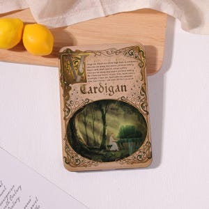 May include: A rectangular tin with a decorative design featuring the word "Cardigan" and an illustration of a person playing a piano in a forest. The tin has a beige background with ornate gold-colored borders and text.