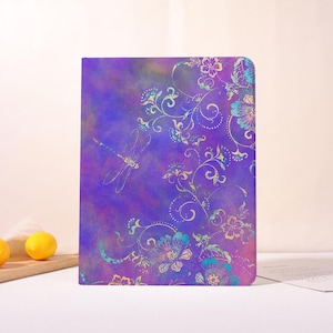 May include: A purple notebook with a dragonfly and floral design. The cover features a watercolor-style background with a dragonfly and floral pattern in teal, gold, and purple. The notebook is rectangular and stands upright.