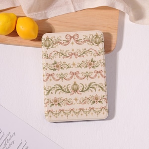May include: A rectangular e-reader case featuring a cream-colored background with a repeating pattern of pink bows, green foliage, small flowers, and gold star accents arranged in horizontal rows across the entire surface.