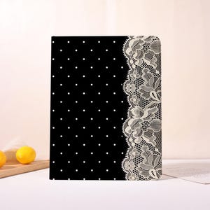 Könnte beinhalten: Ein rechteckiges Notizbuch mit schwarzem Einband, das auf der linken Seite ein weißes Punktemuster und am rechten Rand einen breiten vertikalen Streifen aus detaillierter weißer Spitzenstickerei mit Blumenmotiven aufweist.