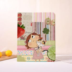 May include: A decorative notebook featuring a cartoon girl hugging a calico cat. The design incorporates strawberries, apples, and a lime slice, with the word "Praise" visible. The background is a patchwork of patterns.