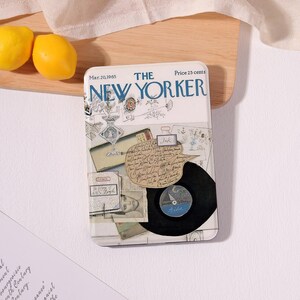 Può includere: Una scatola di metallo rettangolare con una copertina vintage della rivista "The New Yorker" del 20 marzo 1965. Il design include illustrazioni, note manoscritte e un disco in vinile. Due limoni gialli sono visibili sullo sfondo.