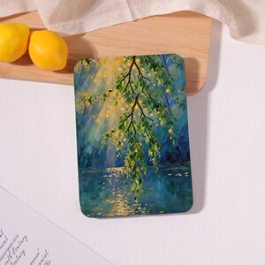 May include: A rectangular e-reader case featuring a printed painting of a tree branch with green leaves and yellow accents hanging over a body of water. The scene depicts sunlight filtering through the branches, creating golden reflections on the blue water.