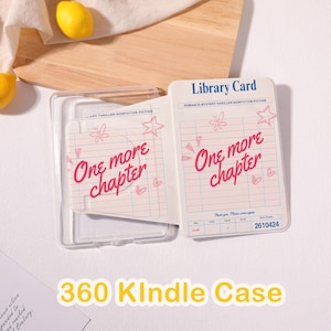 May include: A clear plastic Kindle case featuring a library card design with blue grid lines and pink hand-drawn doodles of stars, hearts, and butterflies. The text reads Library Card, One more chapter, and the number 2610424 in blue ink.