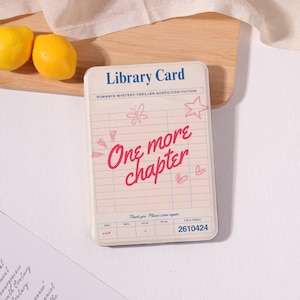 May include: A rectangular card with a cream background featuring the blue text Library Card at the top. The center has a grid with the red cursive text One more chapter, pink hand-drawn doodles of a butterfly, star, and hearts, and the number 2610424.