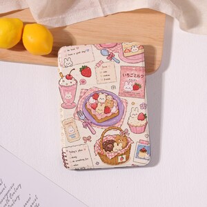 May include: A rectangular notebook with a cream-colored cover featuring a repeating pattern of cartoon bunnies, strawberries, milkshakes, toast, donuts in a basket, and Japanese text on a pink candy bag, alongside handwritten-style notes and lists.