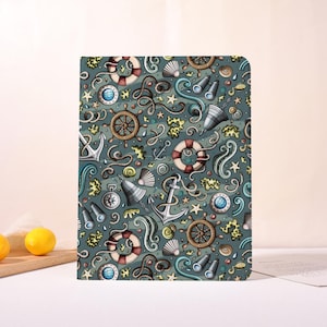 May include: A notebook with a nautical-themed pattern. The design features cartoon illustrations of anchors, ship wheels, life preservers, seashells, and telescopes in shades of blue, white, and brown on a teal background.
