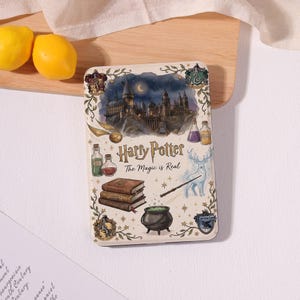 Puede incluir: Lata rectangular con temática de Harry Potter con el castillo de Hogwarts, una snitch dorada, botellas de pociones, libros, un caldero y un ciervo. El texto "Harry Potter The Magic is Real" se muestra en el centro.
