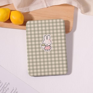 May include: A rectangular electronic reader case featuring a sage green and white gingham check pattern. Centered on the front is a small sticker of a white cartoon rabbit with a pink bow, wearing a pink dress and holding a green drink.
