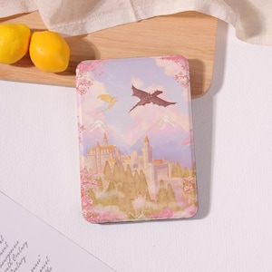 May include: A rectangular e-reader case featuring a pastel-colored illustration of a fantasy castle nestled among trees and mountains. Two dragons fly in the sky above the castle, surrounded by pink cherry blossom branches in a soft, dreamy aesthetic.