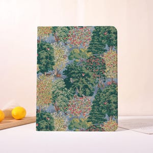 Pode incluir: Um caderno retangular com um padrão de floresta. A capa apresenta uma ilustração detalhada de árvores, folhagens e pequenos animais em tons de verde, vermelho e amarelo sobre um fundo azul. O caderno tem cantos arredondados.