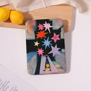 May include: Rectangular art print featuring a person walking down a street with colorful star shapes. The artwork has a dark background with buildings in blue and gold. The person is wearing a red hat and carrying a yellow bag.