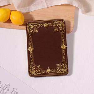 May include: A rectangular, brown leather-bound journal with gold filigree detailing. The cover features an ornate gold border and floral accents. Two yellow lemons rest on a wooden surface next to the journal.