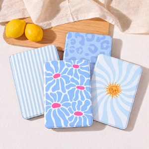May include: Four rectangular metal tins featuring light blue and white designs: one with vertical stripes, one with a leopard print, one with a floral pattern with pink centers, and one with a central sun graphic on a swirling background.