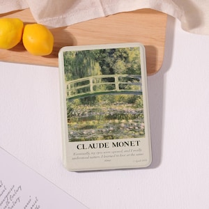 May include: A rectangular metal tin featuring Claude Monet's "Water Lilies" painting. The artwork depicts a green arched bridge over a pond with lily pads. The tin includes the artist's name and a quote. Two yellow lemons sit on a wooden cutting board.