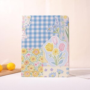 May include: A floral-themed tablet case featuring a patchwork design. The case combines a blue gingham pattern with floral prints in yellow, pink, and blue. A sticker of yellow and pink tulips and daisies is on the front.