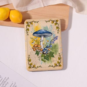 May include: A rectangular e-reader case featuring a central illustration of a large blue mushroom surrounded by smaller brown and blue mushrooms, white daisies, blue bellflowers, and green ivy vines against a background of yellow and blue watercolor splashes.