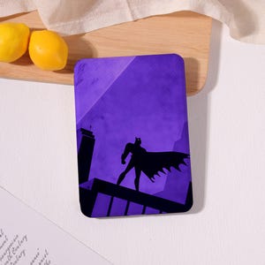 May include: A rectangular protective case for an electronic device featuring a purple background with a black silhouette of a caped figure standing on a rooftop. The design includes sharp geometric shapes representing city buildings under a dark sky.