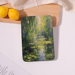 May include: Rectangular coaster featuring a vibrant impressionist painting of a water lily pond with green foliage and reflections. The coaster is on a wooden surface with two yellow lemons.