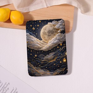 May include: A rectangular tablet case featuring a dark navy blue night sky illustration with a large, pale yellow full moon surrounded by layered, fluffy white clouds. Golden stars hang from thin lines, and a small glowing lantern is visible.