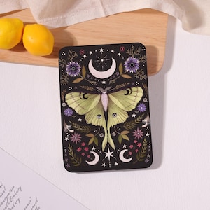 May include: A rectangular e-reader case featuring a black background with a central pale green moth, surrounded by purple flowers, green foliage, white stars, and crescent moons. The design is symmetrical and detailed with fine lines.