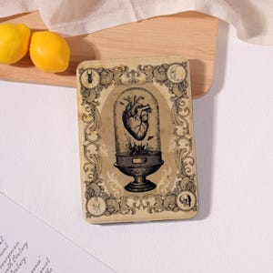 May include: A rectangular wooden plaque featuring a black ink illustration of an anatomical heart inside a glass cloche. The border is decorated with ornate scrollwork, a beetle, a crescent moon, a human figure, and a skull.