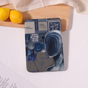 May include: A rectangular e-reader case featuring a blue-toned collage design. The collage includes text such as one day at a time, good things take time, and blue, alongside illustrations of a ship, a moon, and a surreal face with a swirling vortex.