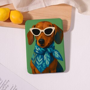 May include: A rectangular tablet case featuring a brown dachshund wearing white-rimmed sunglasses and a blue neckerchief with a light blue leaf pattern, set against a solid green background. The illustration is centered on the front of the case.