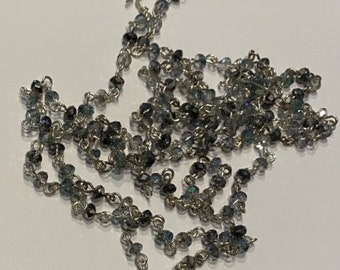 Könnte beinhalten: Eine silberne Kettenkette mit kleinen, facettierten, dunkelblauen und klaren Glasperlen. Die Kette besteht aus kleinen, miteinander verbundenen Metallringen.