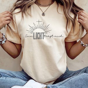 May include: Cream-colored t-shirt featuring a black graphic of a cross, sun rays, and the German text "Sein LICHT tr&auml;gt mich." The shirt has short sleeves and a crew neck.