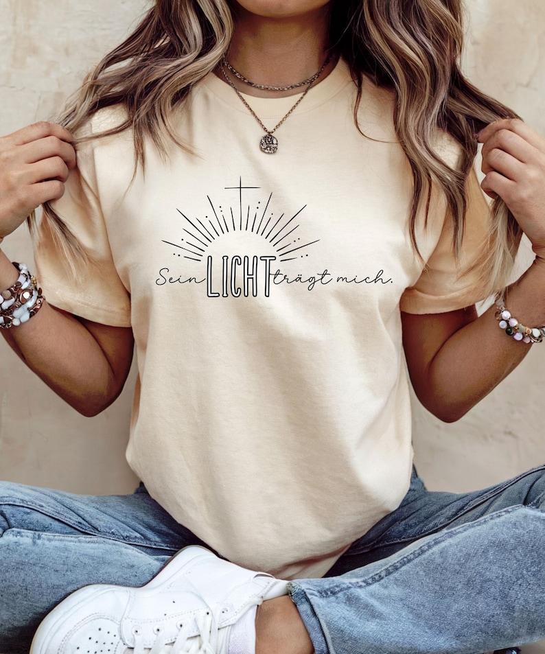 May include: Cream-colored t-shirt featuring a black graphic of a cross, sun rays, and the German text "Sein LICHT tr&auml;gt mich." The shirt has short sleeves and a crew neck.
