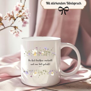 Könnte beinhalten: Weißer Keramikbecher mit floralem Design in Lila, Gelb und Weiß. Der Becher trägt den Text "Du bist kostbar, wertvoll und von Gott geliebt!" und "Mit stärkendem Bibelspruch".