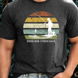 Könnte beinhalten: Dunkelgraues T-Shirt mit einer Grafik eines Anglers in einem Sonnenuntergangsdesign. Der Sonnenuntergang besteht aus horizontalen Streifen in Orange, Gelb, Grün und Blau. Der Text "KÖDER REIN. STRESS RAUS." ist darunter gedruckt.