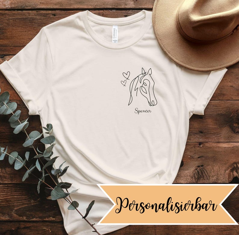 K&ouml;nnte beinhalten: Cremefarbenes T-Shirt mit einer minimalistischen schwarzen Strichzeichnung eines Pferdekopfes und zwei Herzen. Der Name "Spencer" steht darunter. Ein beiger Hut ist im Hintergrund zu sehen. Das Shirt ist aus weichem Material.