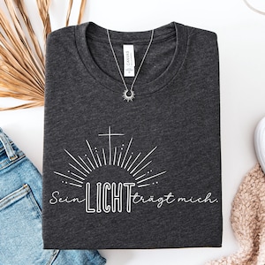 May include: Dark gray t-shirt with the text "Sein LICHT tr&auml;gt mich" in white, featuring a cross and sunburst design. A silver necklace with a sun pendant is on the shirt. The shirt is folded on a white surface.