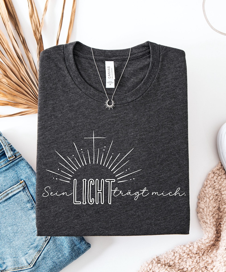 May include: Dark gray t-shirt with the text "Sein LICHT tr&auml;gt mich" in white, featuring a cross and sunburst design. A silver necklace with a sun pendant is on the shirt. The shirt is folded on a white surface.