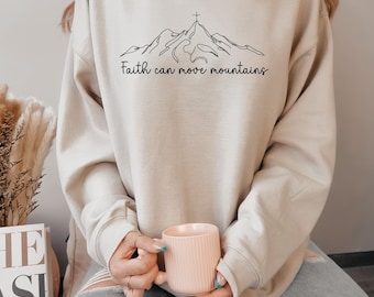 May include: A beige sweatshirt with a mountain graphic and the text "Faith can move mountains" in black. The sweatshirt is paired with light-wash jeans. A pink mug is held in the hands.