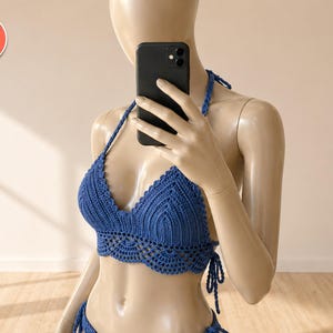 May include: A blue crochet bikini top with a halter neck and tie closures. The top features a scalloped edge and a detailed crochet pattern. The image also shows a matching bottom, and the logo "Sun-Kissed & Covered".