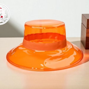 May include: An orange, transparent sun hat with a wide brim and a band around the crown. The hat is made of a shiny material and sits on a light-colored surface. A wooden box is in the background.
